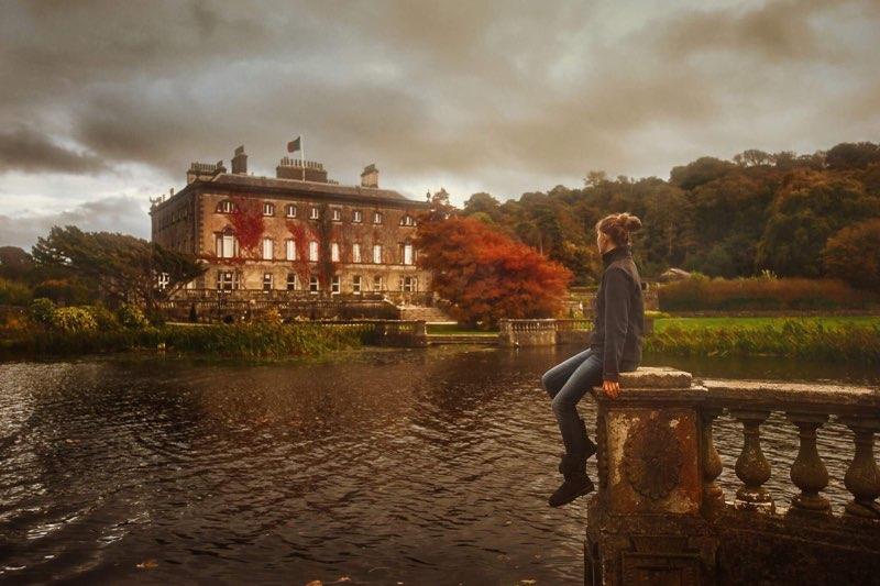 Young woman sitting on a stone fence  next to a pond overlooking the Westport House on a fall overcast day.