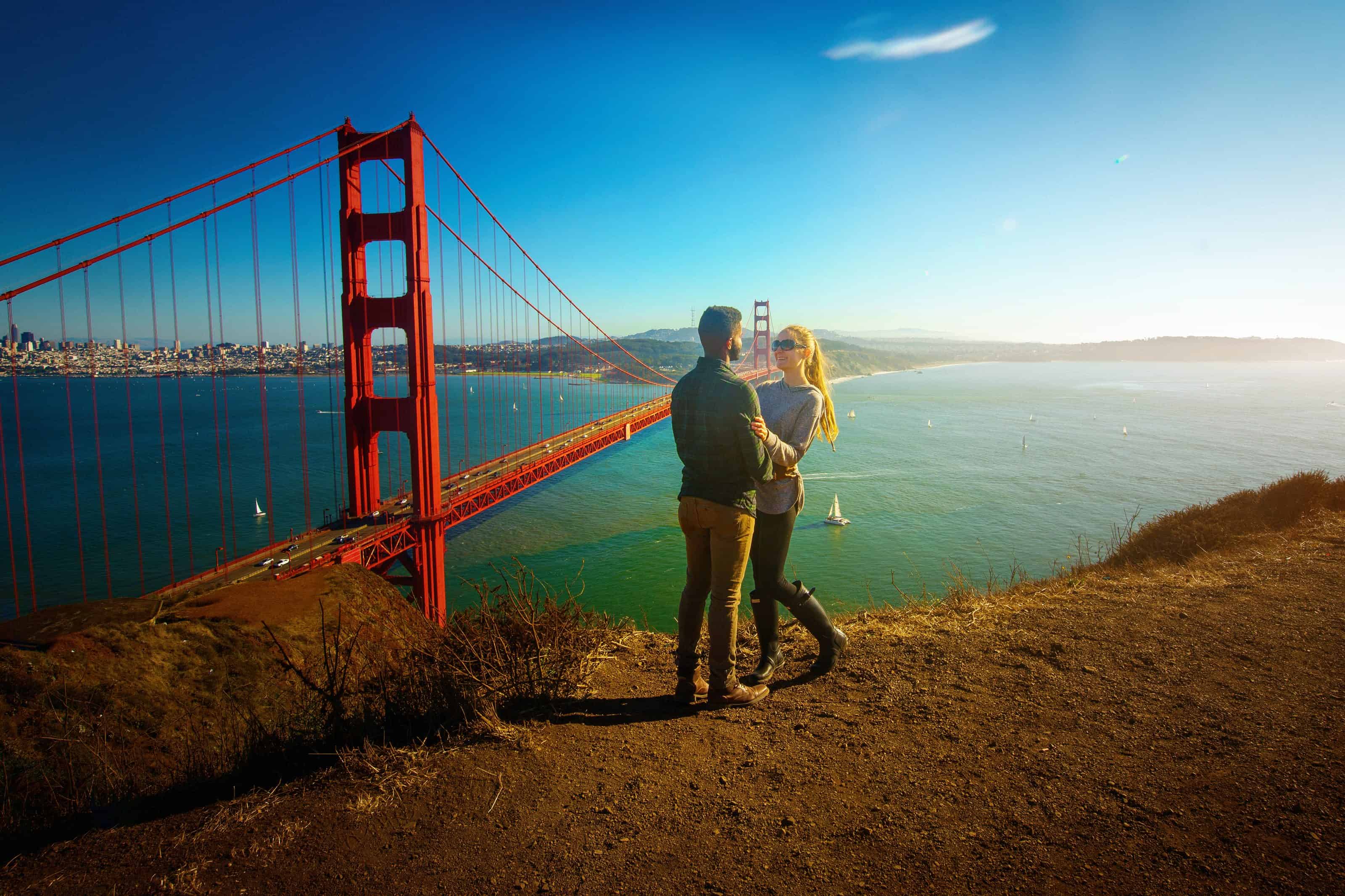 Couple standing on a cliff overlooking the Golden Gate Bridge at golden hour.