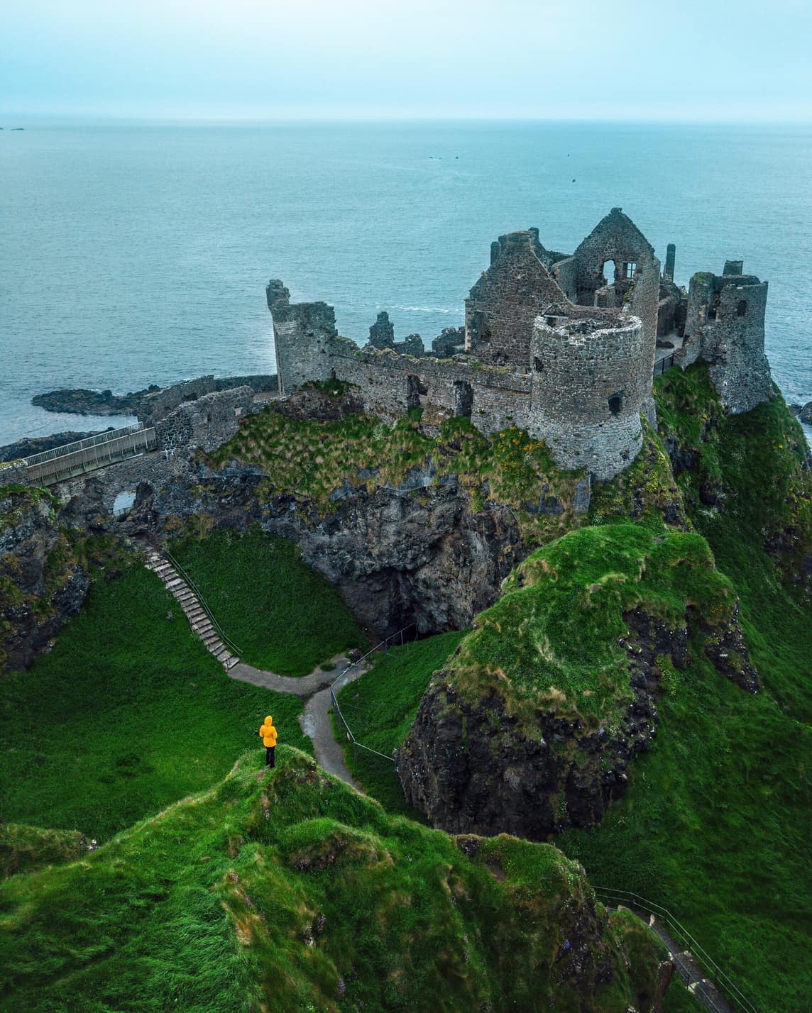 Figure in a yellow raincoat standing on a grass hill overlooking the stone ruins of the Dunluce Castle on the coast during an Ireland road trip.