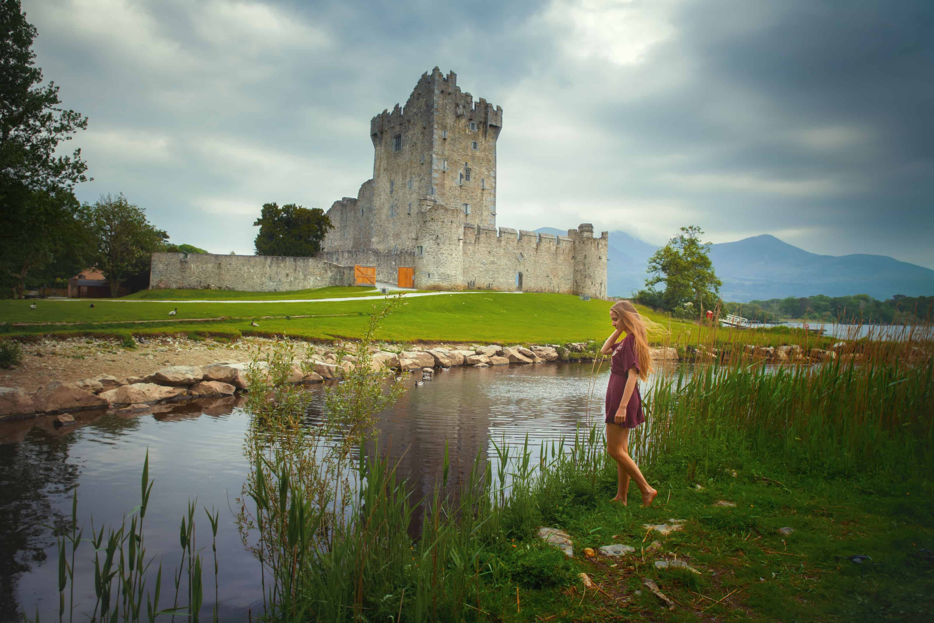 Young woman standing next to a river and the stone Ross Castle on a cloudy day.