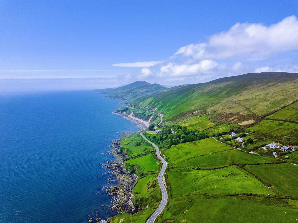 Aerial view of a winding coastal, Ireland road with the blue ocean on one side and green fields on the other side.