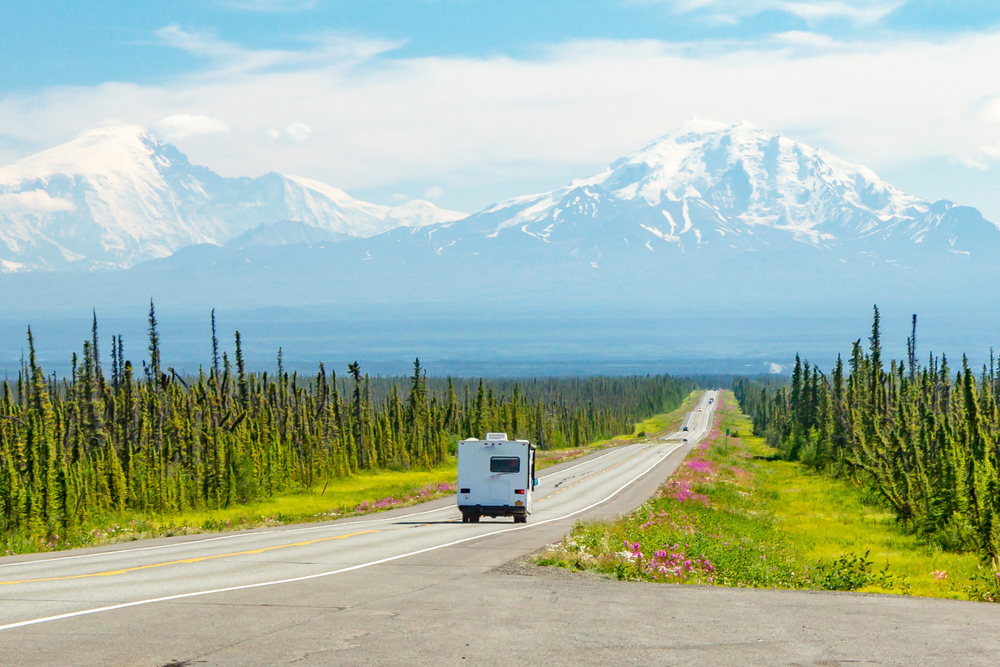 An RV on a road heading towards the mountains among flowers and green trees on an Alaska road trip.