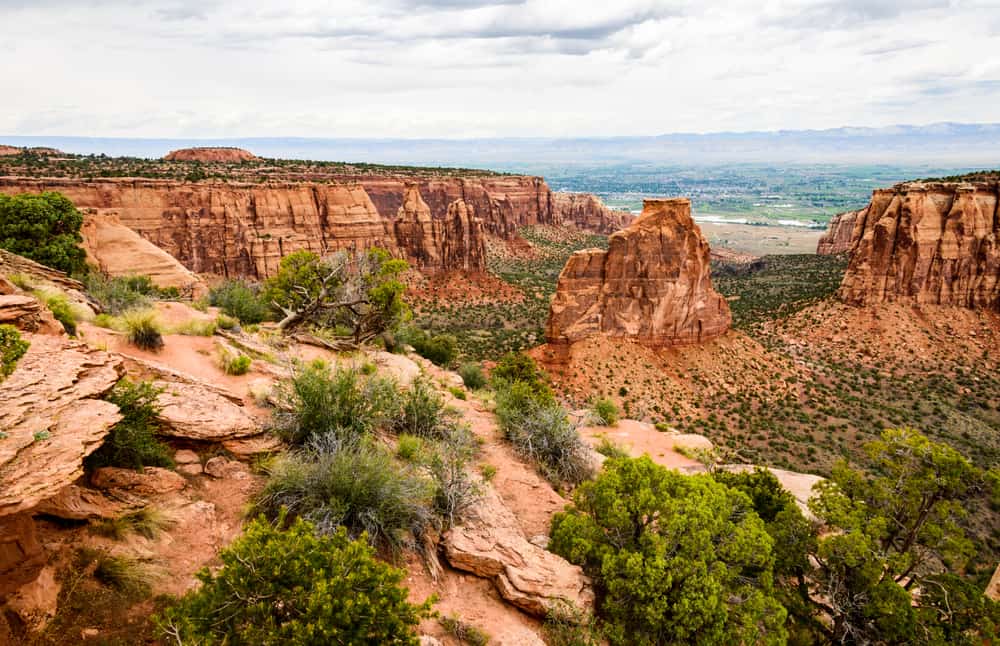 Wide photo of red rock formations at Colorado National Monument with green trees in the foreground.