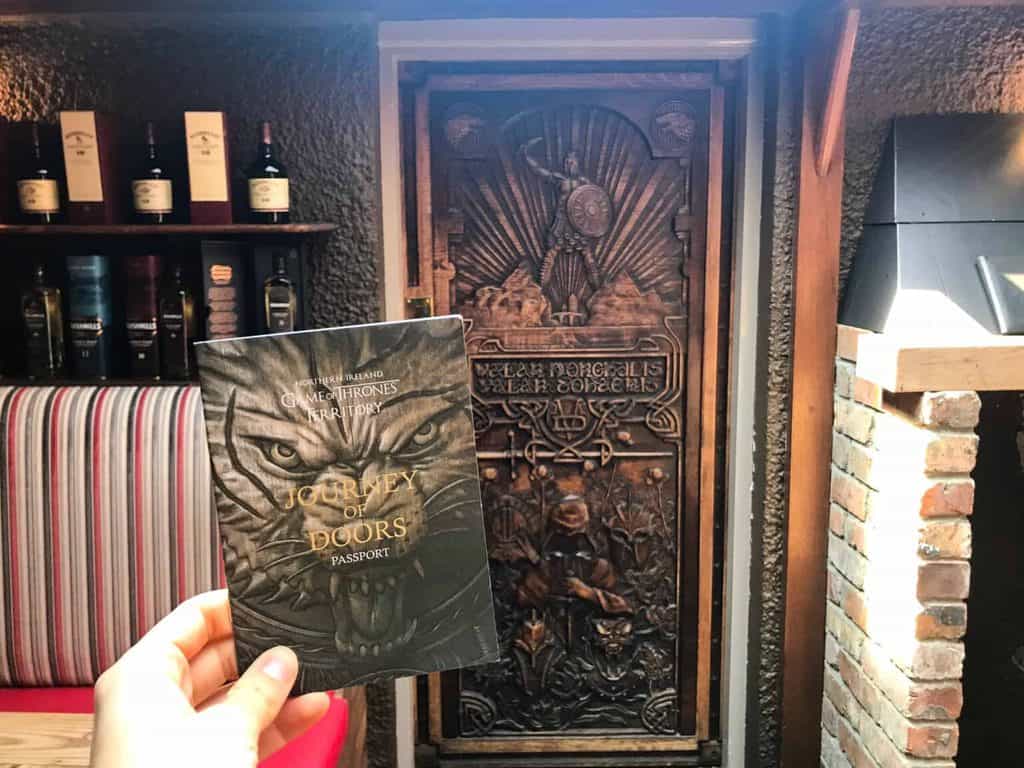 Hand holding a pamphlet of the Game of Thrones doors in front of a wooden door carves with Game of Thrones scenes.