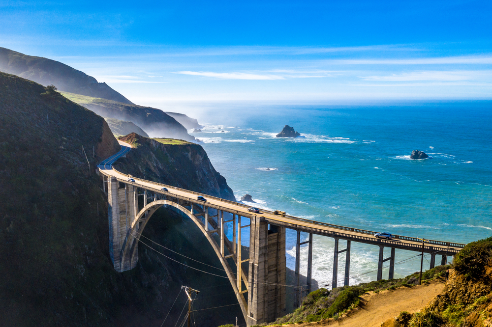 A side aerial view of the famous Bixby Bridge on the Big Sur, a road in California. The bridge connects two mountains and behind it you can see the pacific ocean with a few rock formations in it.  