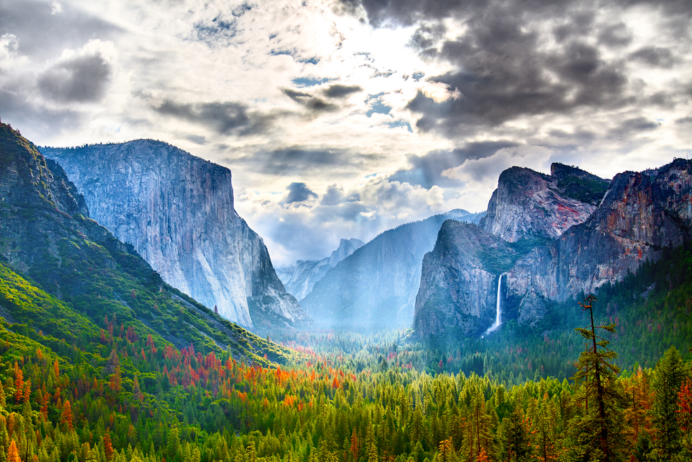 A gorgeous landscape at Yosemite National Park. There are trees in the valley in between large mountains. On one mountain you can see a waterfall on the side of it. It is cloudy but there is sun shinning through the clouds. 