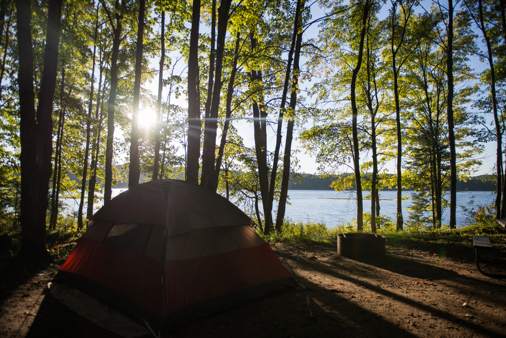 A tent hidden behind trees peers out over the water and rising sun in Michigan. Despite it not being an ocean, the Great Lakes are one of the top areas for the best beach camping in the USA. 
