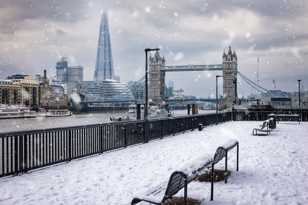 Snowy and cloudy day over the River Thames and benches with the Tower Bridge and Shard building in the background.