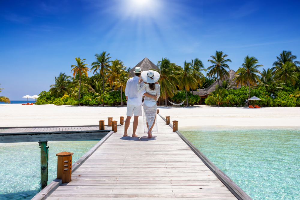 A beautiful traveler couple in white clothing stands on a wooden pier and enjoys the view to a tropical paradise island at the Maldives, Indian Ocean