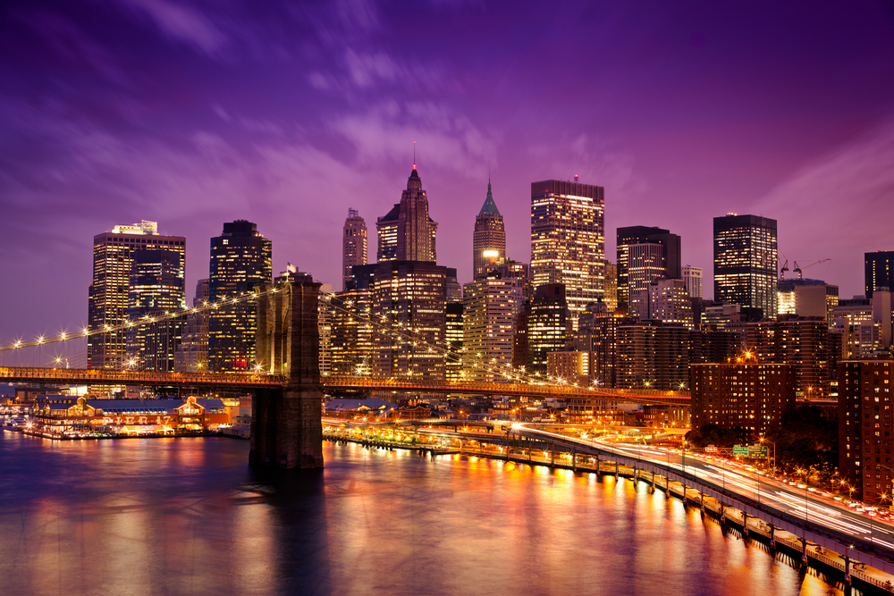 Skyline of downtown New York with a bridge in the foreground. the lights are all on in the skyscrapers. 