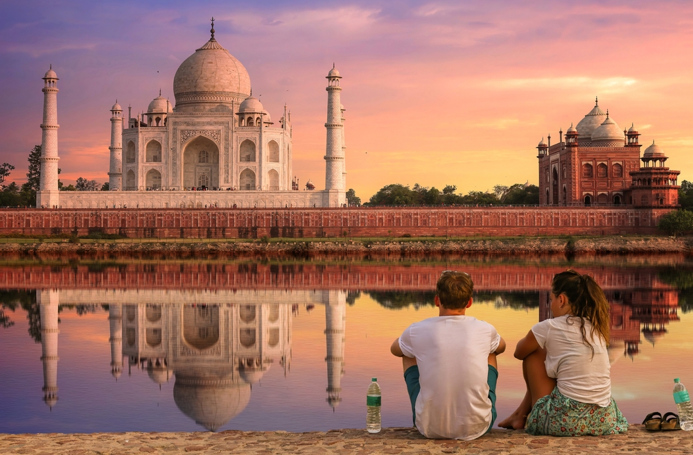 Taj Mahal Agra at sunset with moody sky and water reflection enjoyed by tourist couple sitting at Mehtab Bagh beside river Yamuna. The article is about  Best Vacation Spots For Couples. 