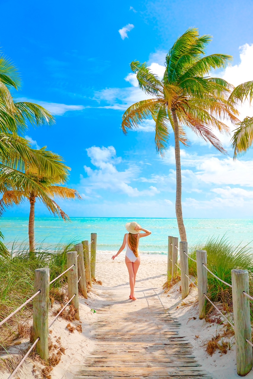 Me wearing a white bathing suit and wide brimmed straw hat looking at a beach in the Florida Keys. The Water is a beautiful azure and palm trees are on either side. A great 30th birthday destination. 