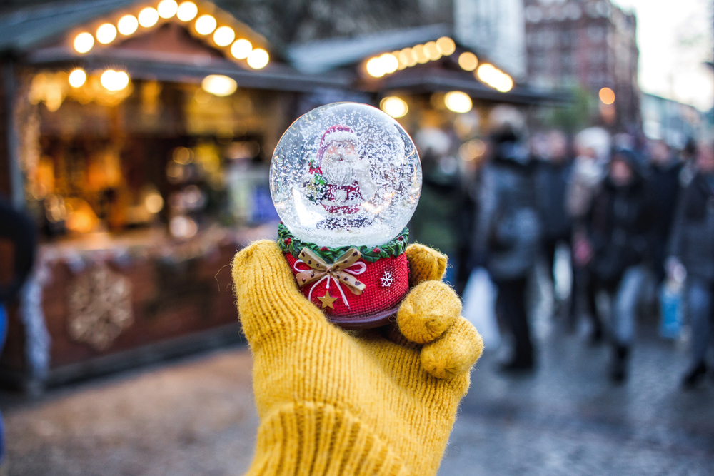 Manchester, one of the best Christmas cities in Europe, features handmade gifts and food, as seen in this photo that shows a gloved hand that holds a snow globe in front of blurred Christmas market huts. 