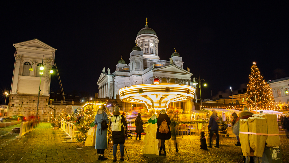 A photo of Helsinki shows people walking around the the Christmas Market in the square, with threes and a spinning carousel! 