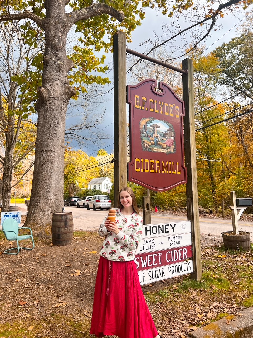 A woman in a red skirt and a mushroom sweater holds onto cider and donuts in front of a Cidermill, which is a great foodie stop! 