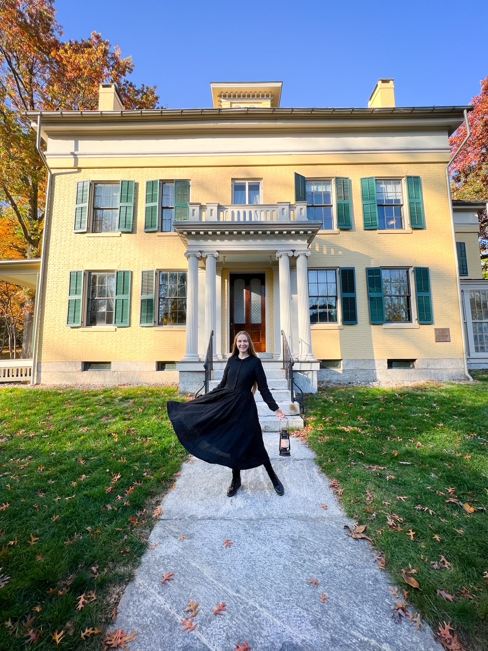 A woman in a black dress holding a lantern, stands in front of the home of Emily Dickinson, whose house is two story with green shutters and yellow paint. 