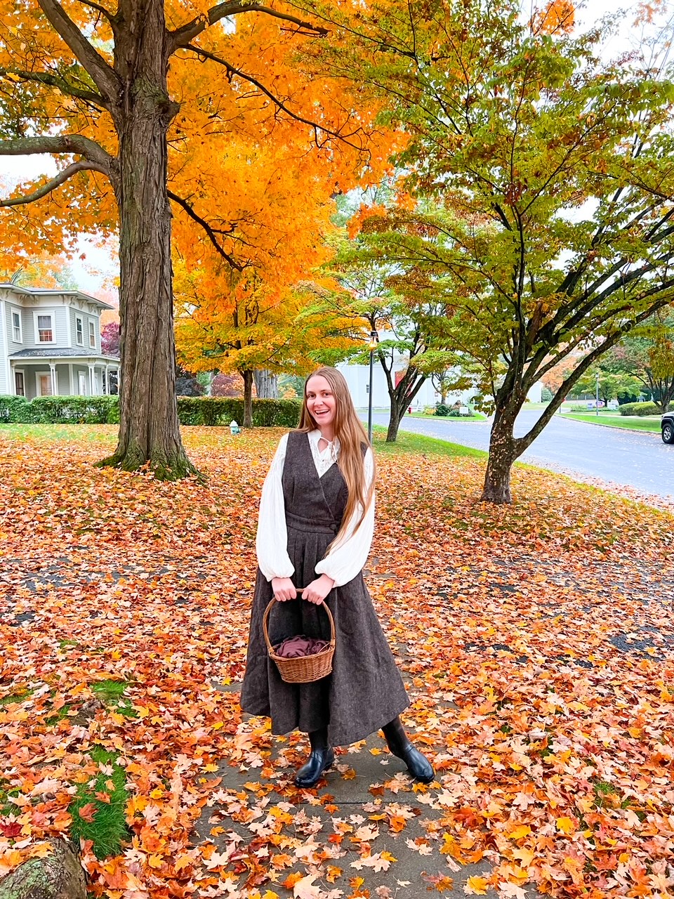 A women dawning a long grey dress and a white-long sleeved shirt underneath calls back to historic times while she holds a straw basket and stands on a street that is full of leaves during a Connecticut fall foliage road trip. 