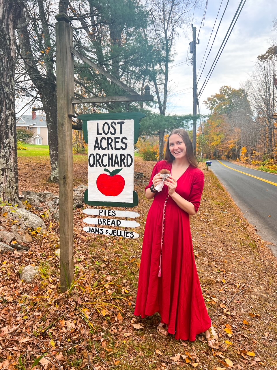 A woman in a red dress holds an apple cider donut in front of the Lost Acres Orchard  sign, which is full of fun, local fall things to do.  