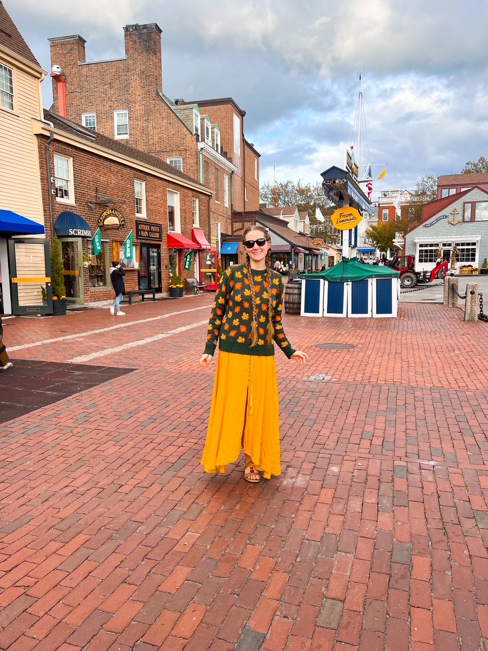 A woman in a yellow skirt and a fall-leaf sweater stands in front of the cute shops of downtown mystic during a Connecticut fall foliage road trip. 