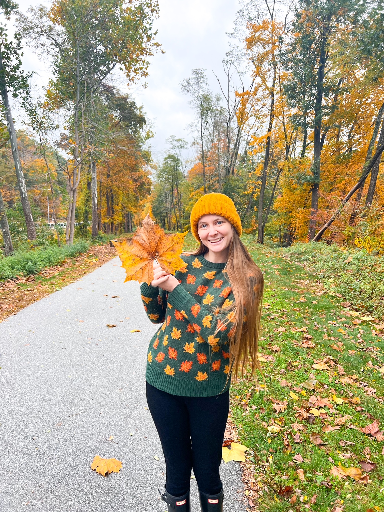 A woman in a yellow hat and a green-leaf sweater holds a huge yellow leaf next to her head while grinning. 