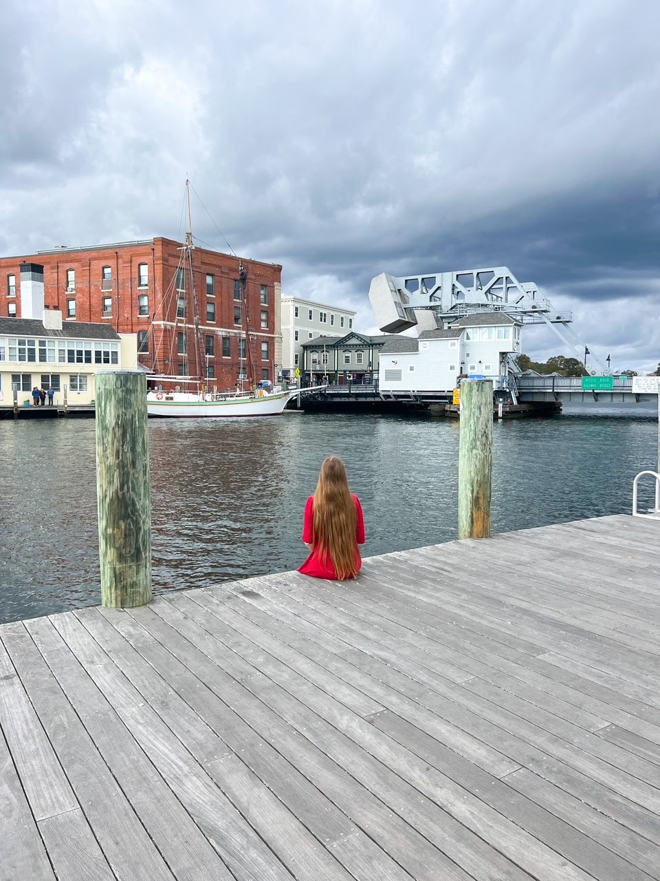 A woman on a pier looks over the water and at the draw bridge of downtown Mystic. Her hair is down and she wears a bright red dress that contrasts the blue of the water and town. 