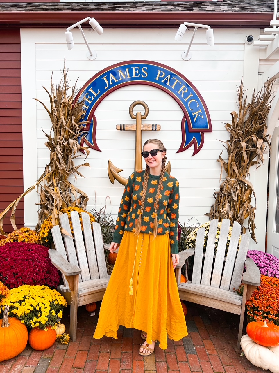 A women smiles in front of the MJP store, wearing a classic fall-inspired sweater from here: the yellow and orange leaves on the sweater mass the foliage of a Connecticut fall foliage road trip. 