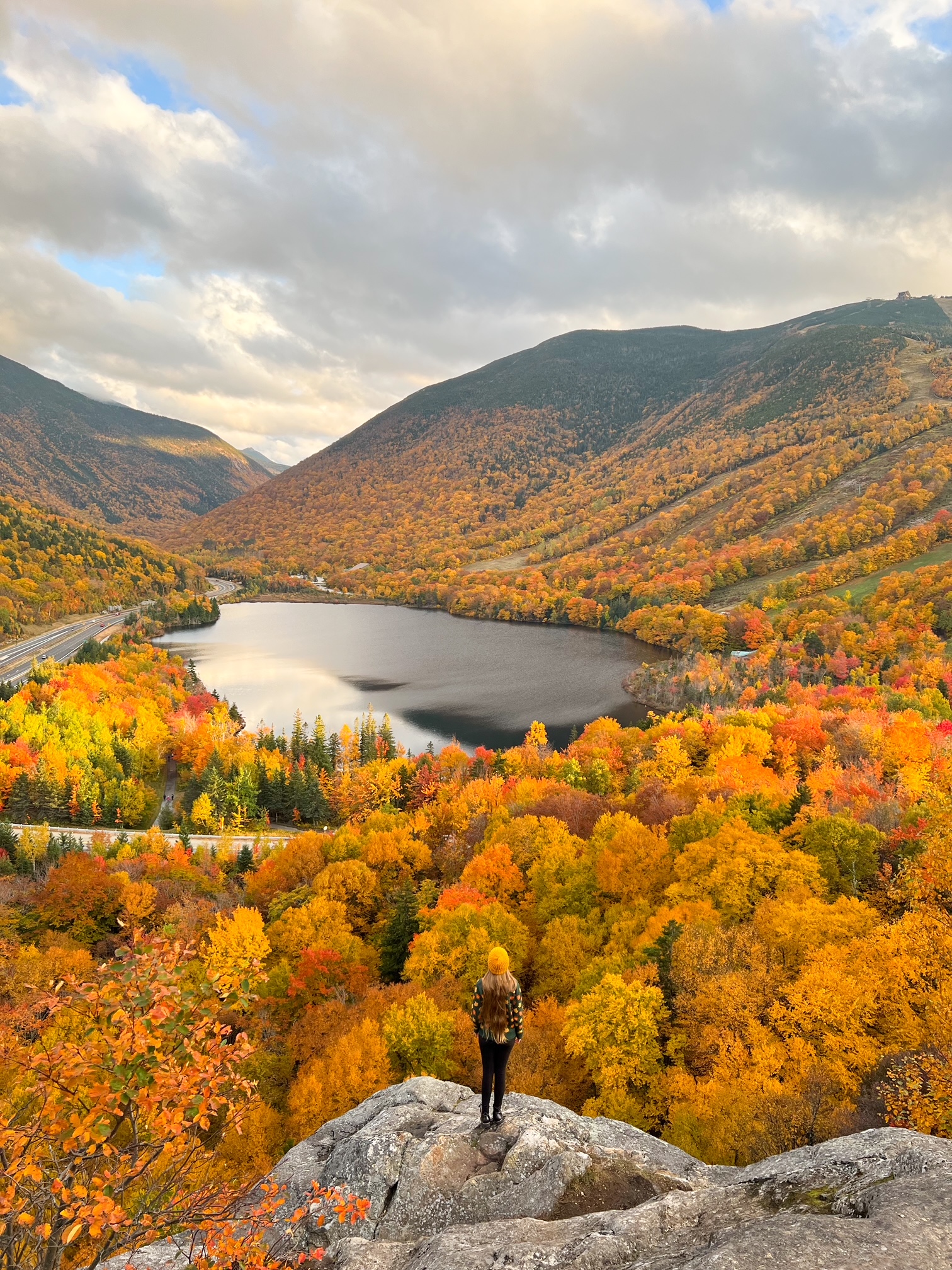 A women in a yellow hat and green sweater stands at the look out of Artist's Bluff: the orange trees scatter around a beautiful lake, making this a must see during your New England Fall Foliage Road Trip Itinerary. 