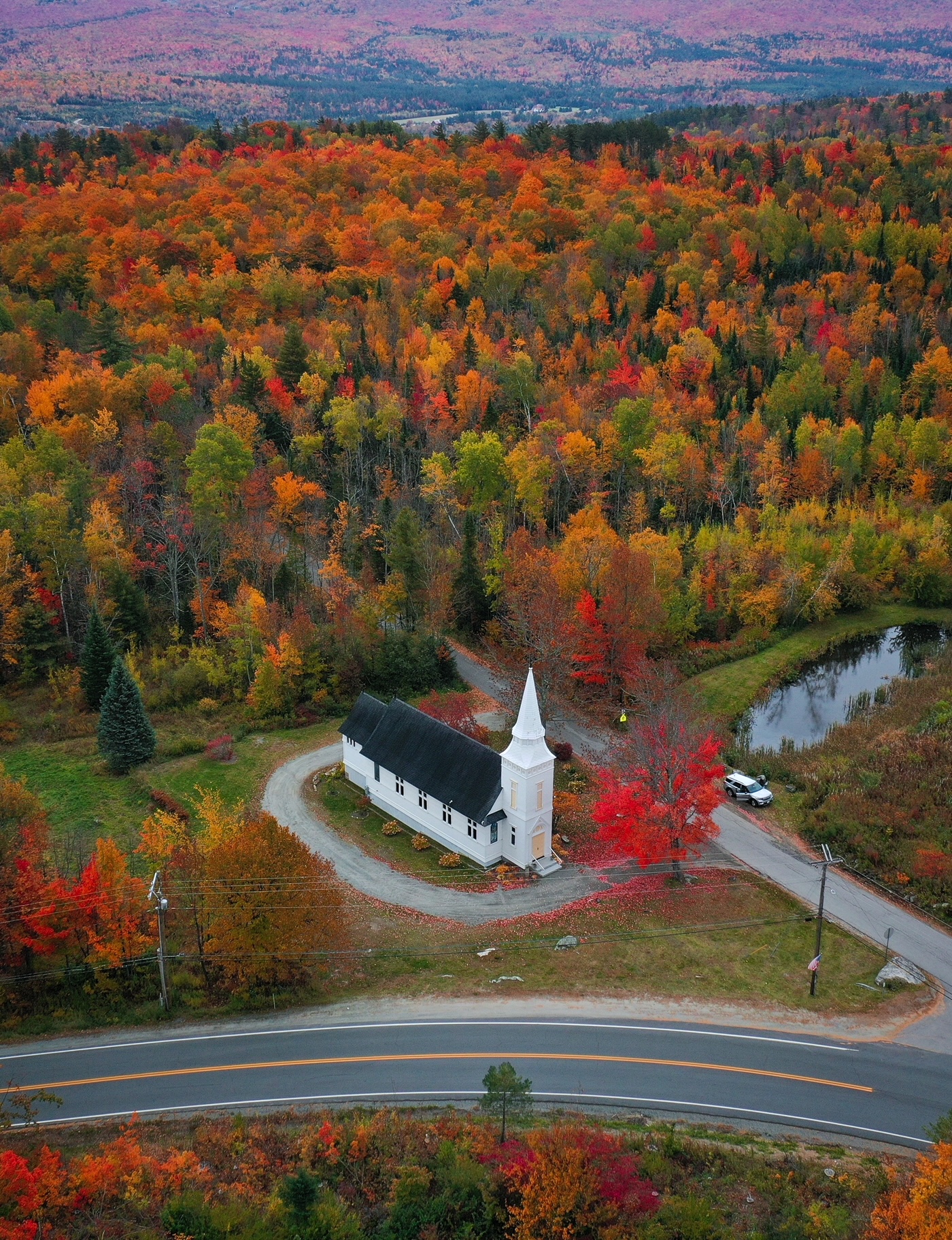 A drone show shows a white chapel at the bend of a road, surrounded by bright red trees. 