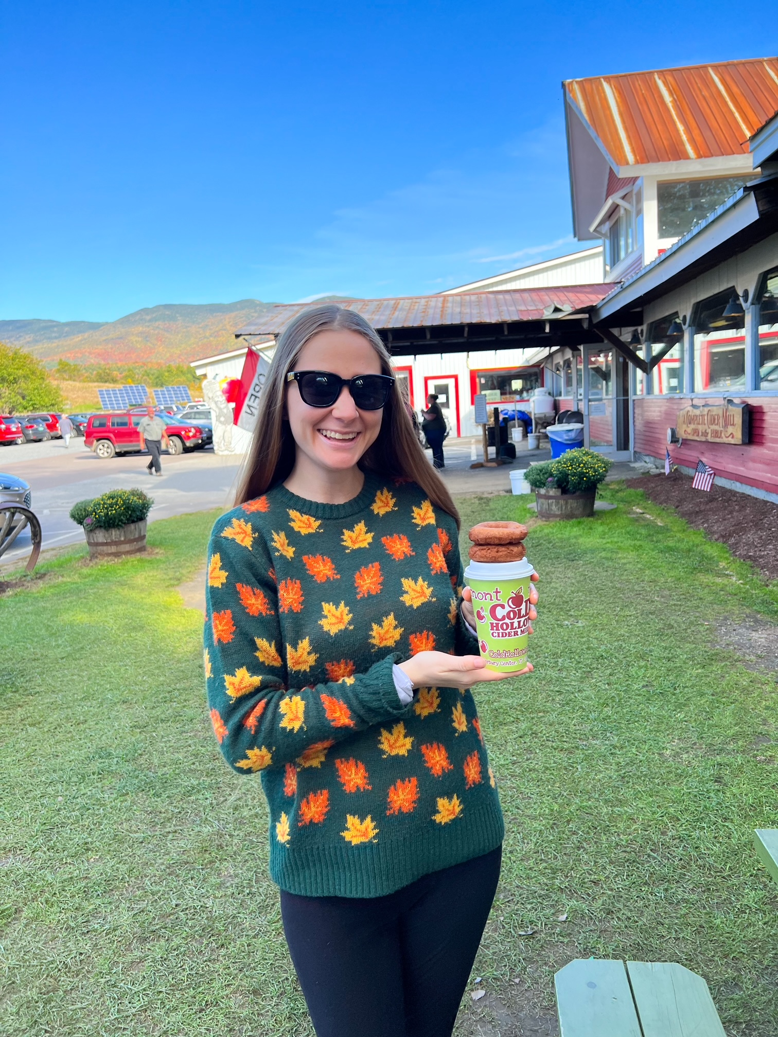 A women in a fall-themed-leaf sweater holds a cider cup and two donuts at Cold Hollow Cider Mill, a must see stop for your  New England Fall Foliage Road Trip Itinerary. 