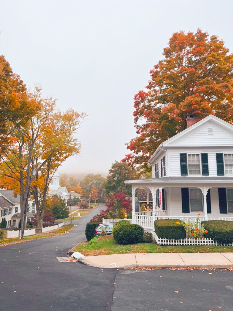 A white home sits on the corner of a street with a white, wrapped around porch and stunning fall foliage. You'll see lots of places like this during your New England Fall Foliage Road Trip Itinerary. 