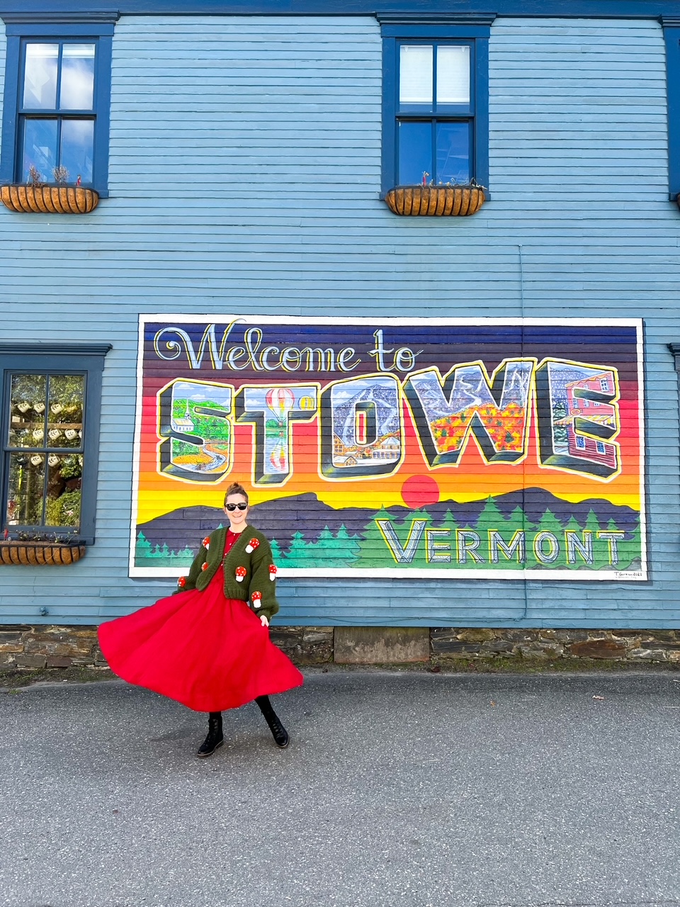 A women in a red skirt and mushroom sweater spins in front of the Welcome to Stowe Vermont sign, that features mountains and a sunset. This is a place to stop add to your New England Fall Foliage Road Trip Itinerary.