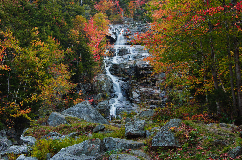 A larger waterfall with darker rocks and powerful waves of water sits in the middle of red-lined trees at Sabbaday Falls, which is a must stop for your New England Fall Foliage Road Trip Itinerary. 