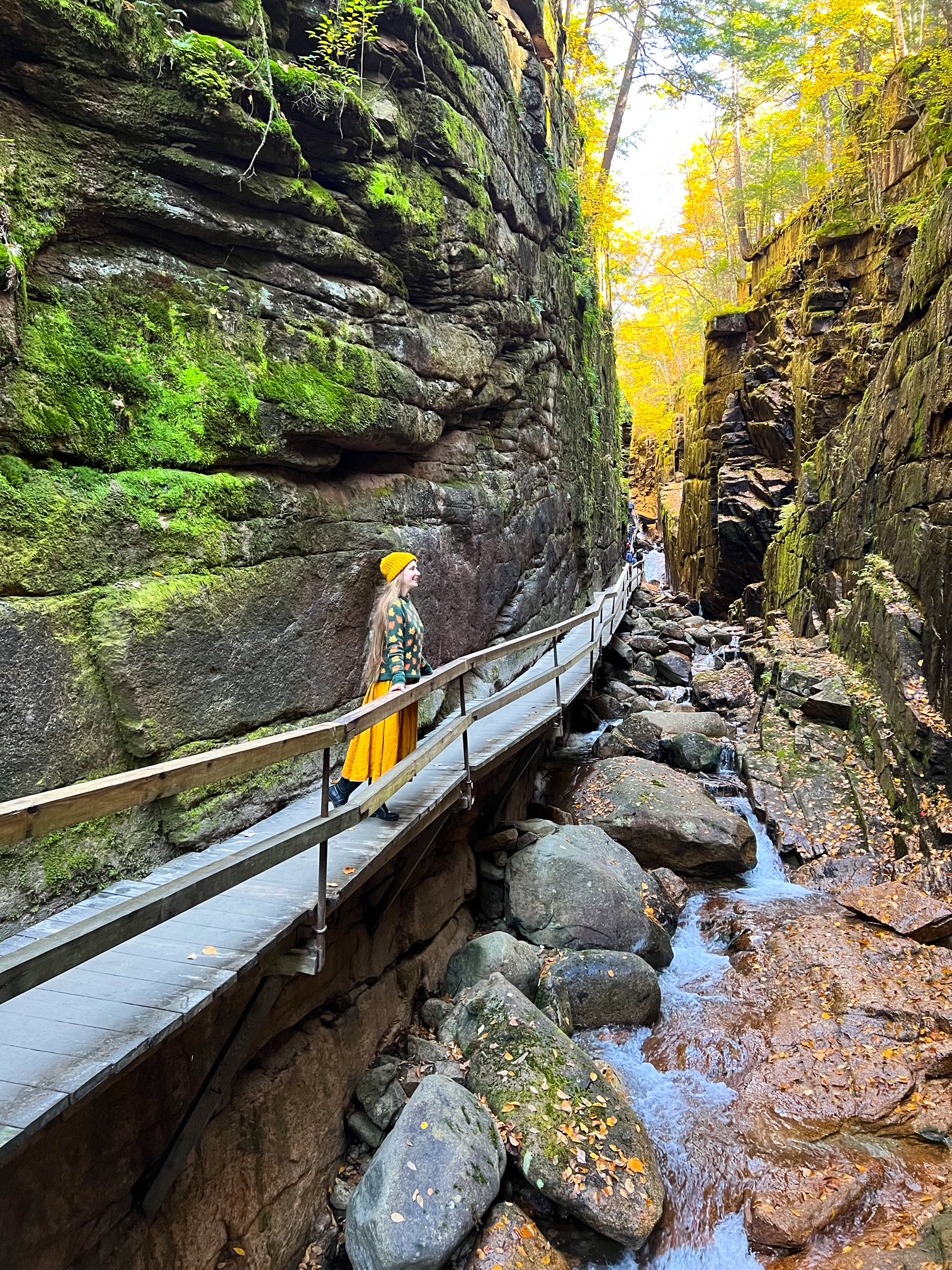 A women enjoys the views of Flume Gorge from below: in her yellow skirt and hat she stares at the rick formations that divide the gorge making this a must see during your New England Fall Foliage Road Trip Itinerary.