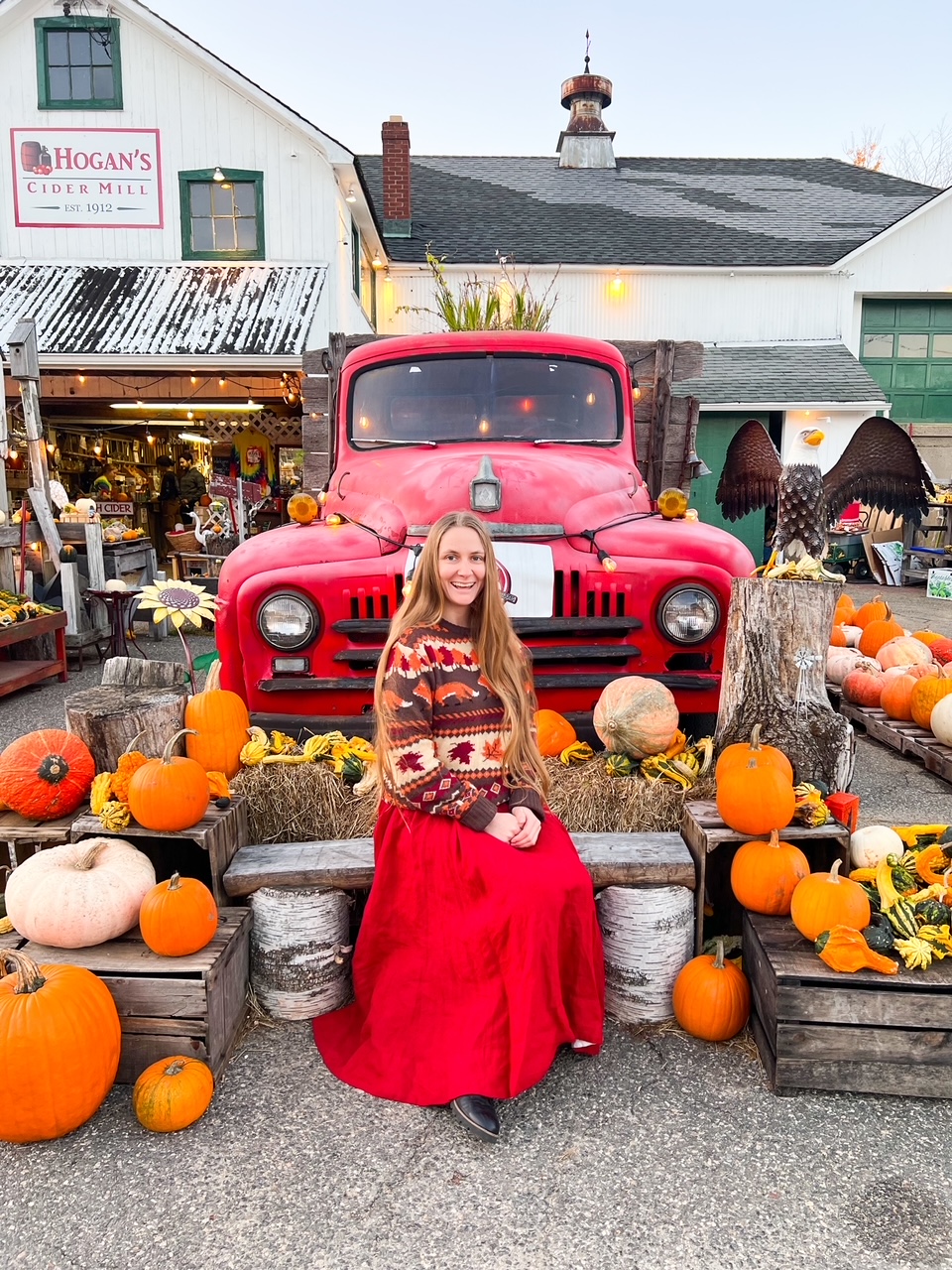 A girl sits in front of a red truck with pumpkins for a cute photo op at Hogan's Cider Mill, a place you have to add to your New England Fall Foliage Road Trip Itinerary. 