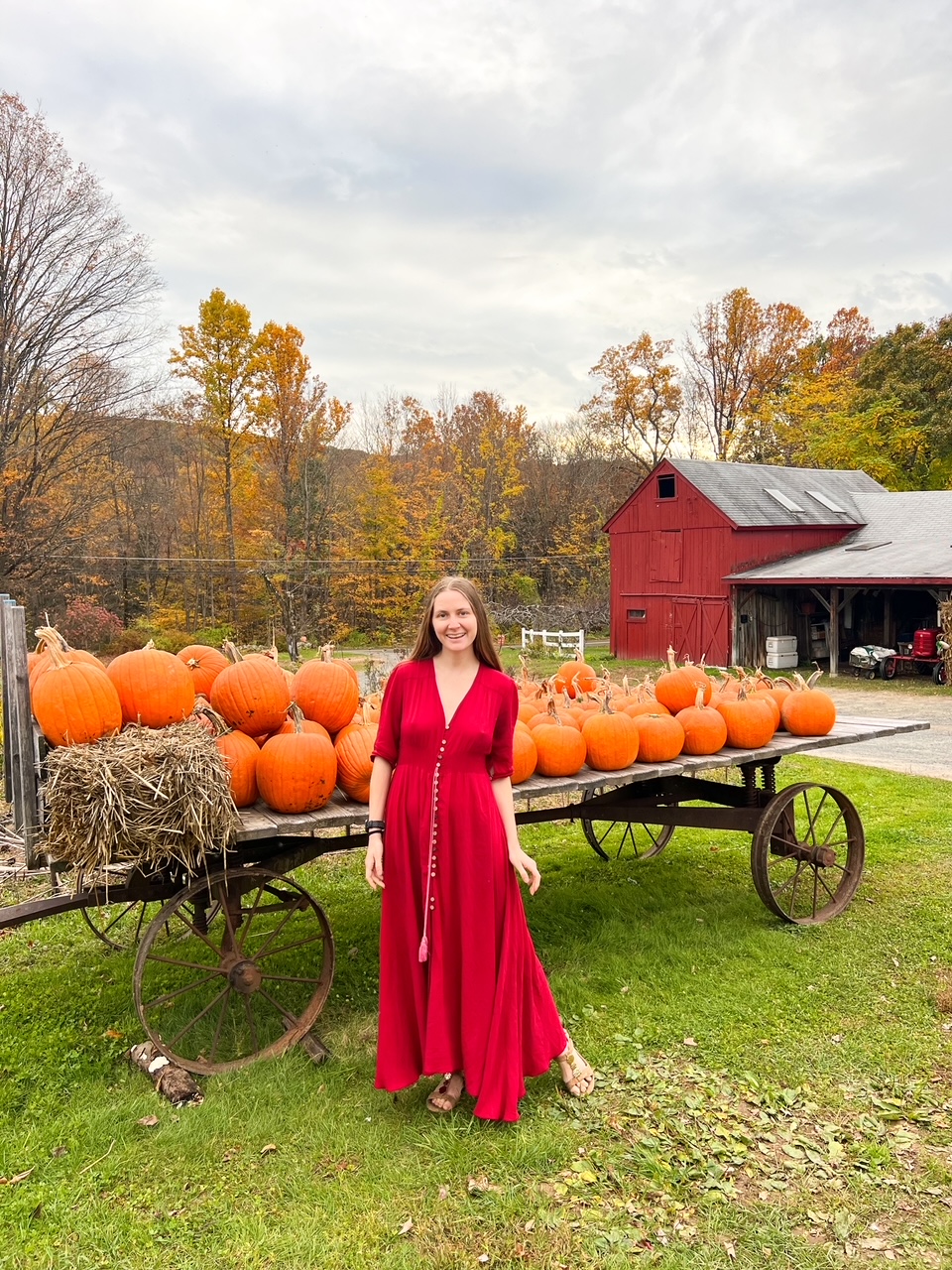 A women in a red dress stands in front of a pile of orange pumpkins: there are so many fun fall things to do during your New England Fall Foliage Road Trip Itinerary. 