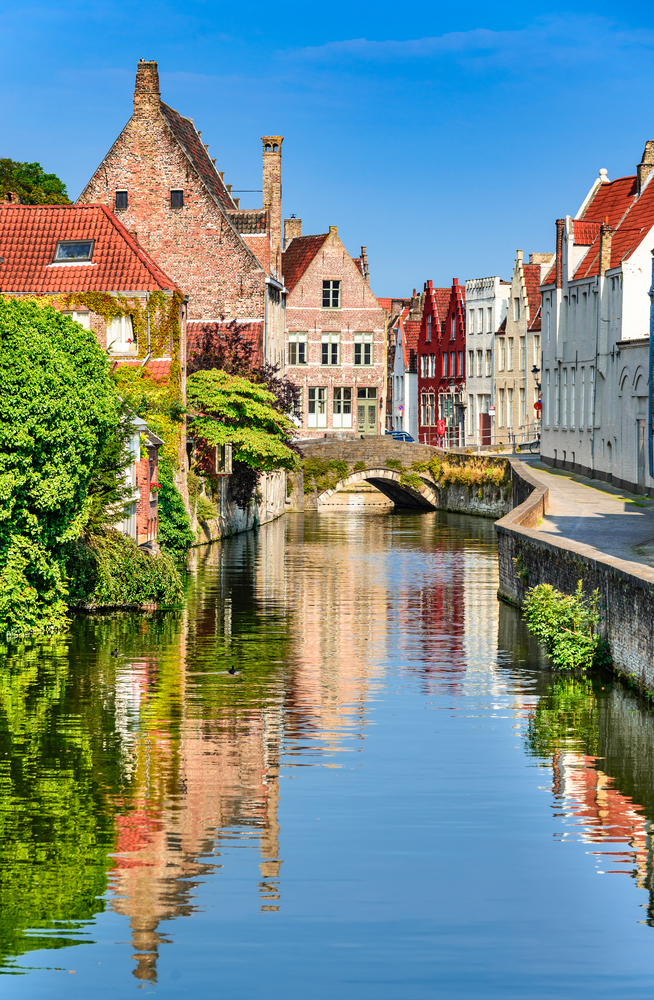 a canal through bruges, there is an arched bridge over the water and vegetation on both sides of the canal