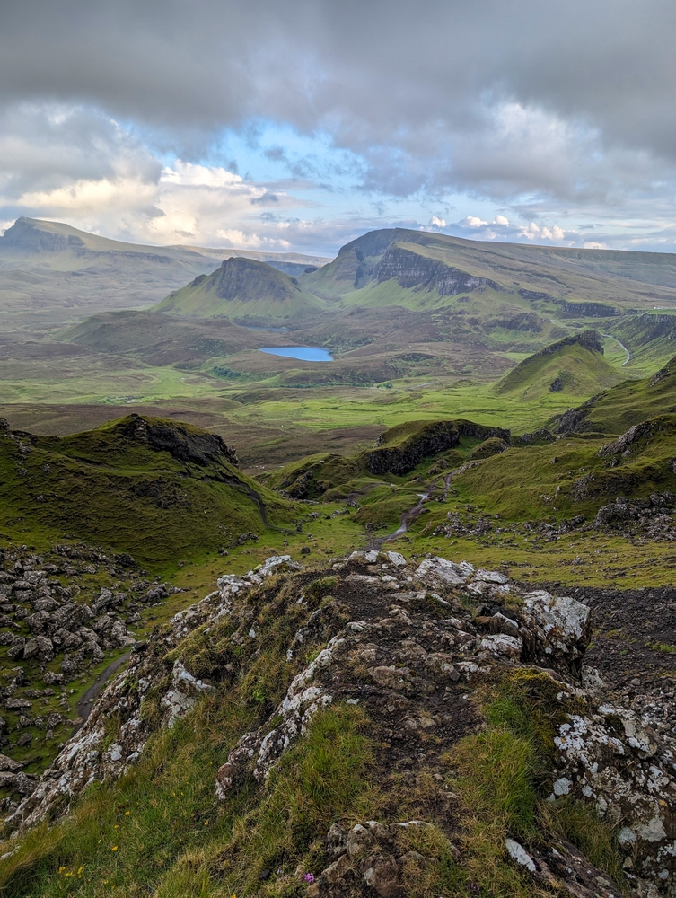 rocky mountains and rolling hills with green grass on them on a mostly cloudy day, a lake is shown in the distance