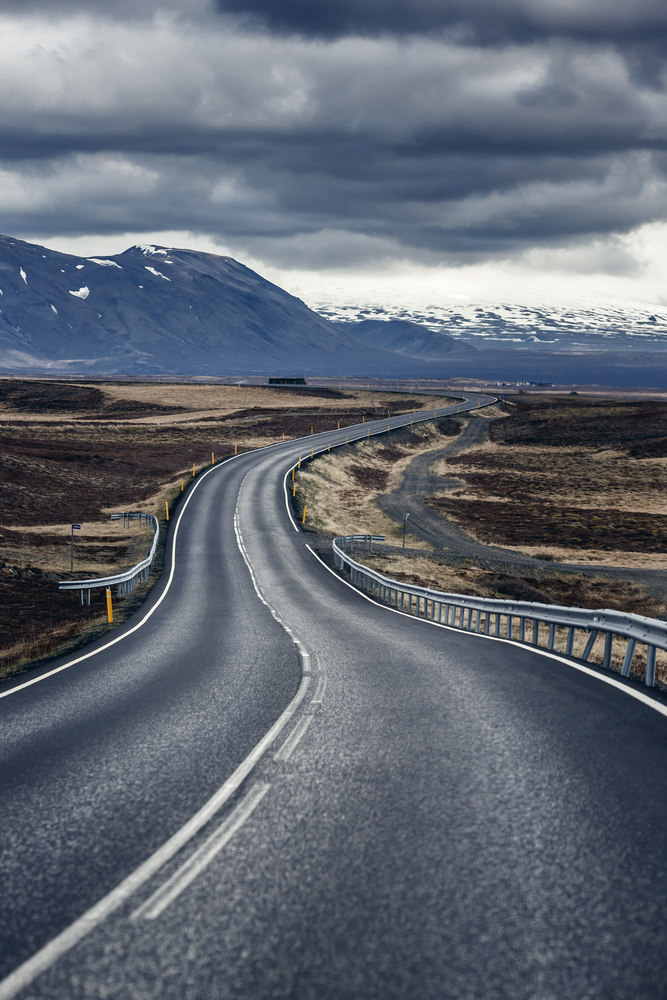 a winding road in iceland on a cloudy day, there are snow covered mountains in the background