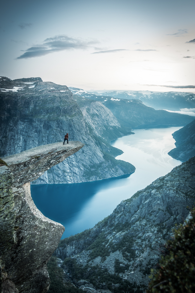 a person stands on a rock plateau overlooking a fjord, there are snow covered mountains in the background