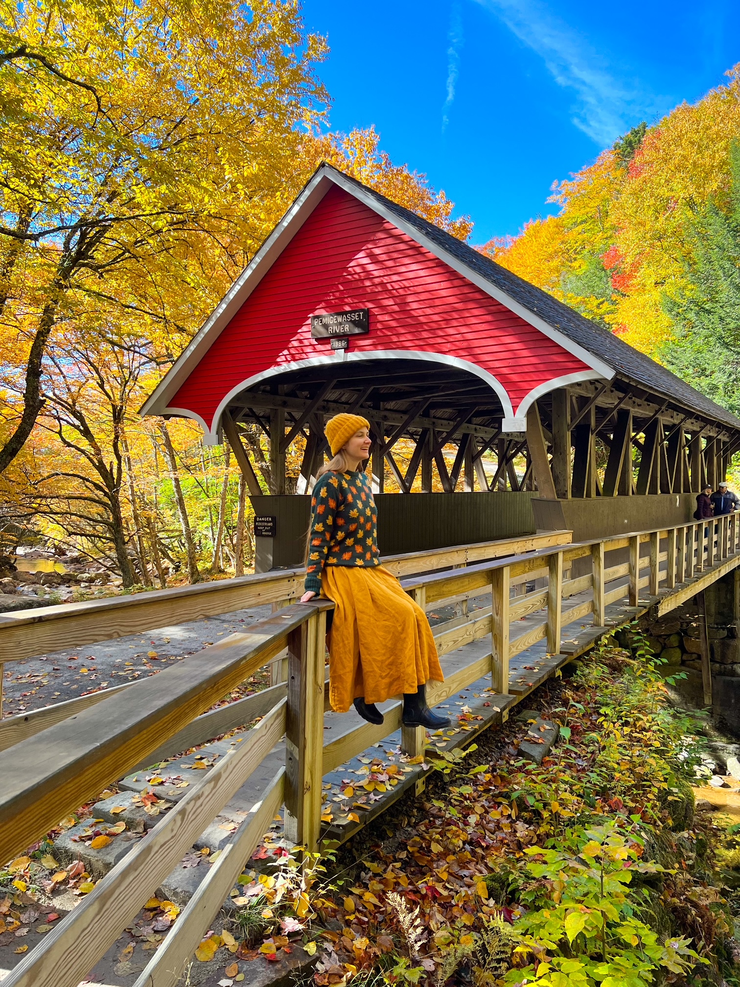 A woman in a yellow-orange hat and skirt sits on the edge of a railing by a covered bridge, enjoying the views of the Flume Gorge at Fraconcia Notch State Park. 