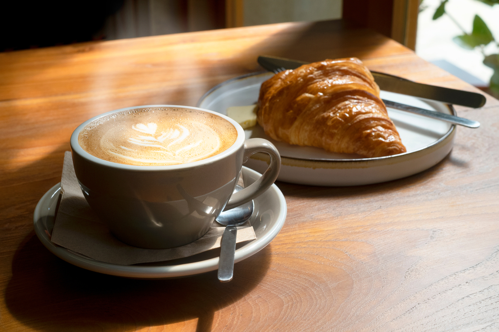 A steaming latte with a design made from the cream sits next to a butter croissant on a table. 