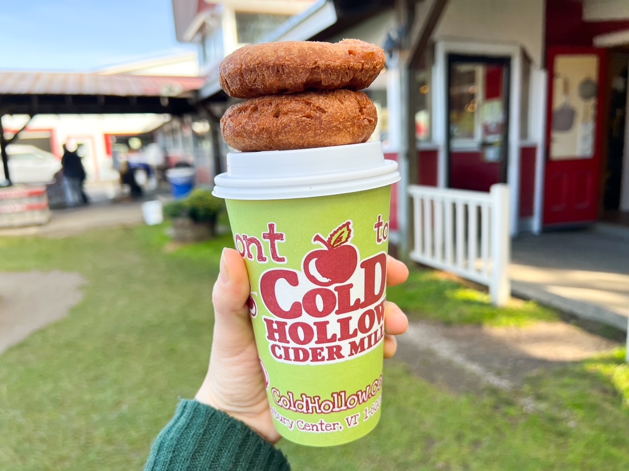 A hand holds a to-go cup of cider from Cold Hollow Cider Mill, and two donuts it a top the cup! 