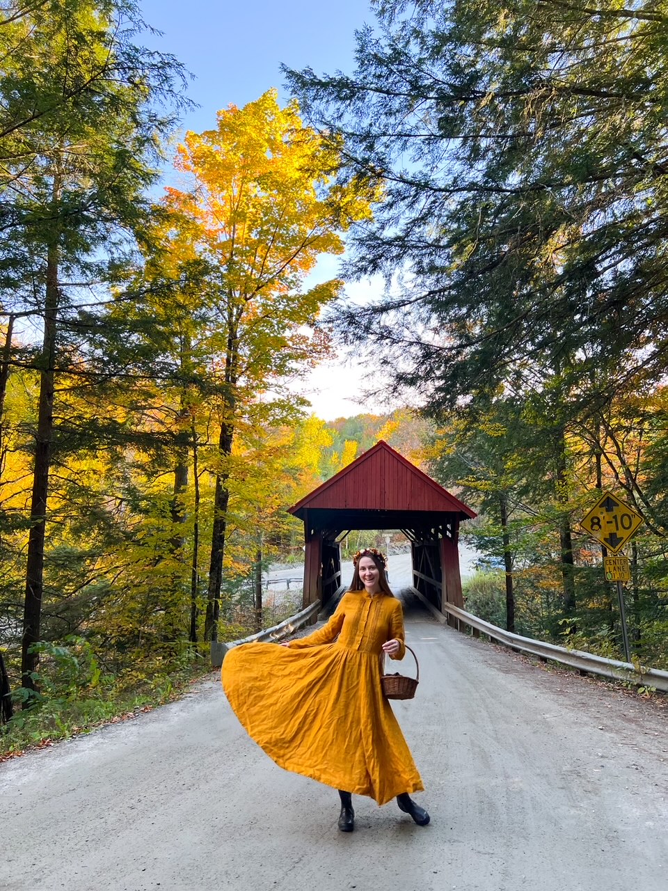 A woman in a yellow dress who carries a small basket on her arm stands in front of a covered bridge on a small road, surrounded by trees. 