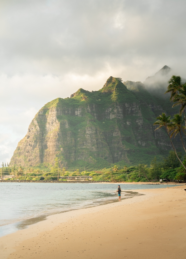 Beautiful sunset tropical beach in Oahu, Hawaii. You can see the beach with a mountain in the background. There is one man stood in the water. 