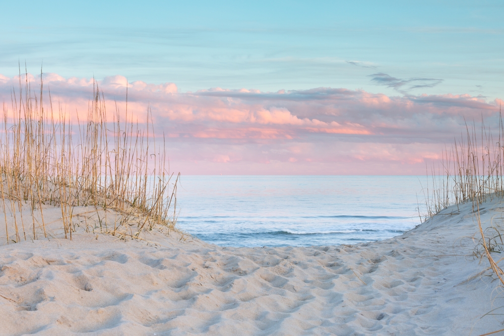 Sandy path of footprints leading through the dunes and grasses to the beach and the Atlantic Ocean under a pastel sky of pink and blue at sunset on the Outer Banks of North Carolina.