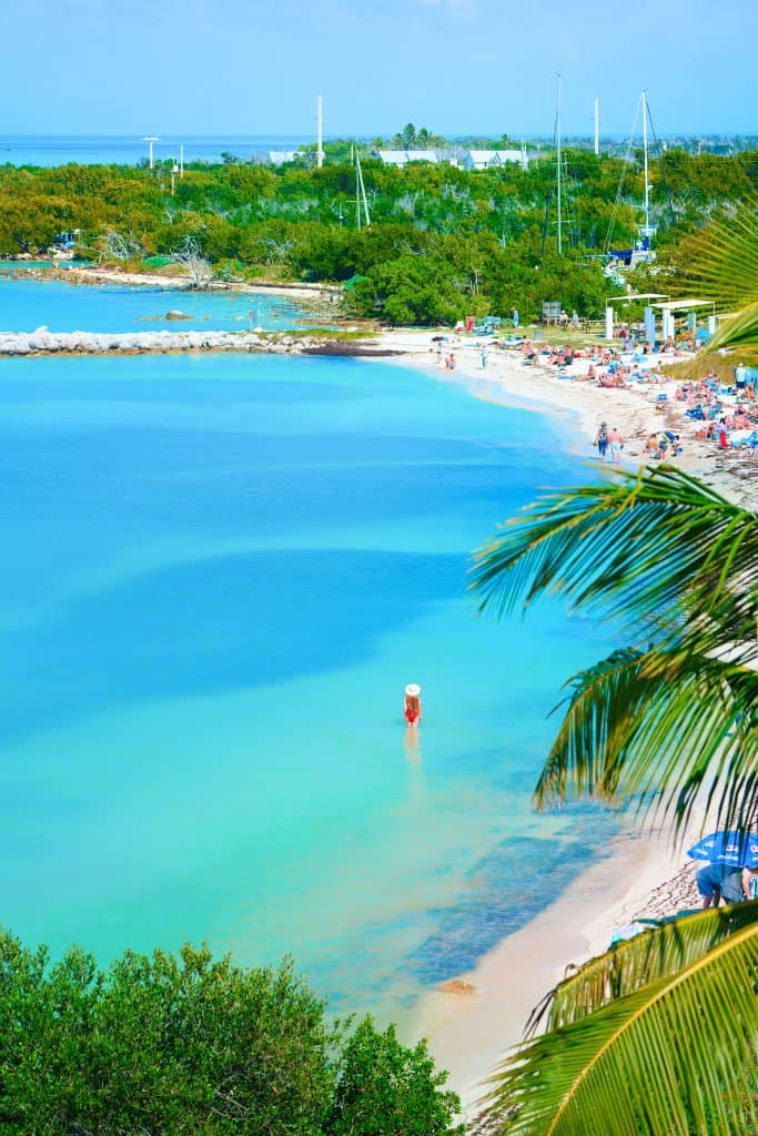 View of Bahia Honda you can see the beach and trees. There is a girl in a red swimsuit and hat is in the foreground of the water. 