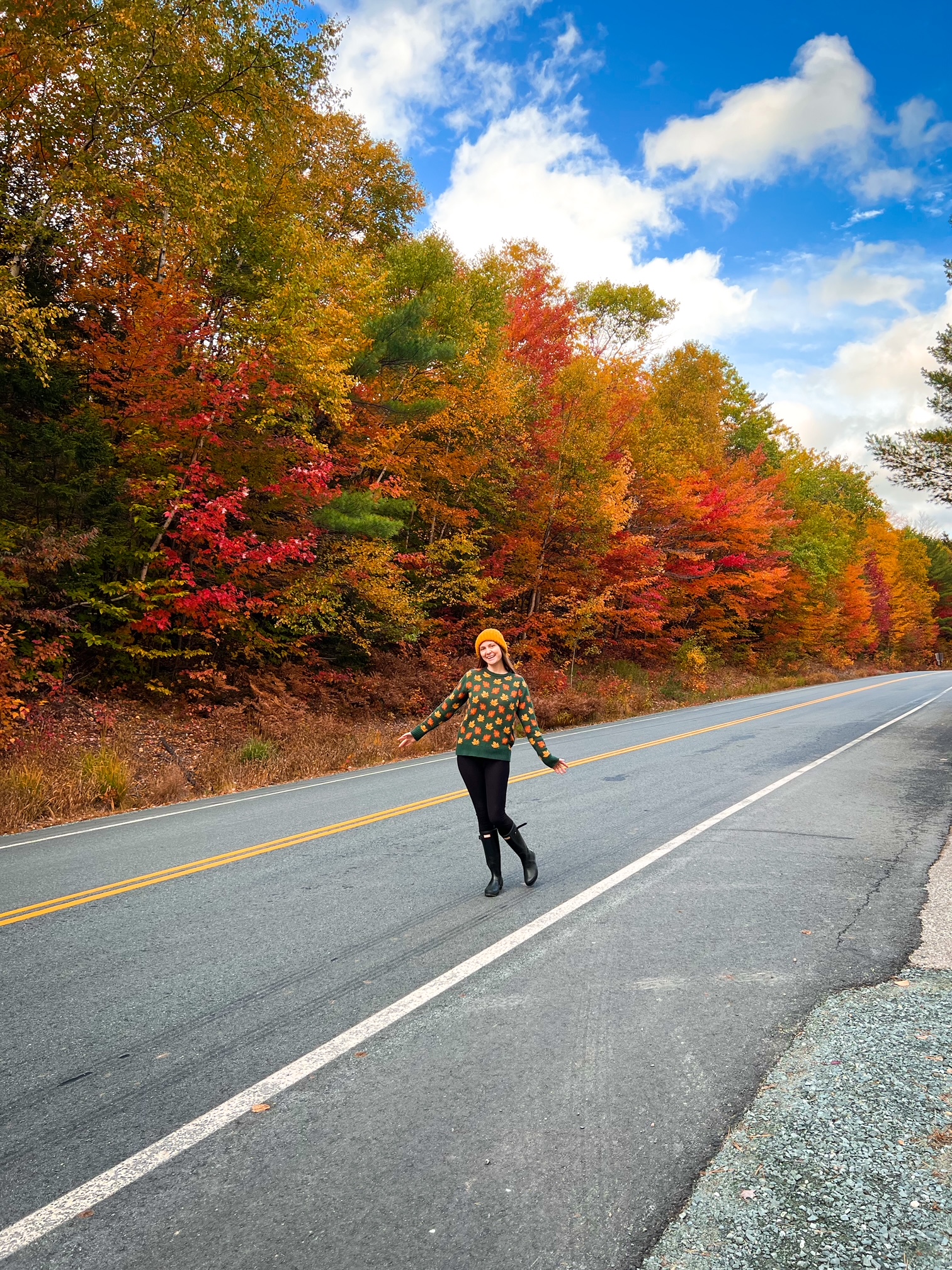 A woman in a sweater with fall colored leaves matches the fall foliage behind her as she stands in the middle of a New England Road. 