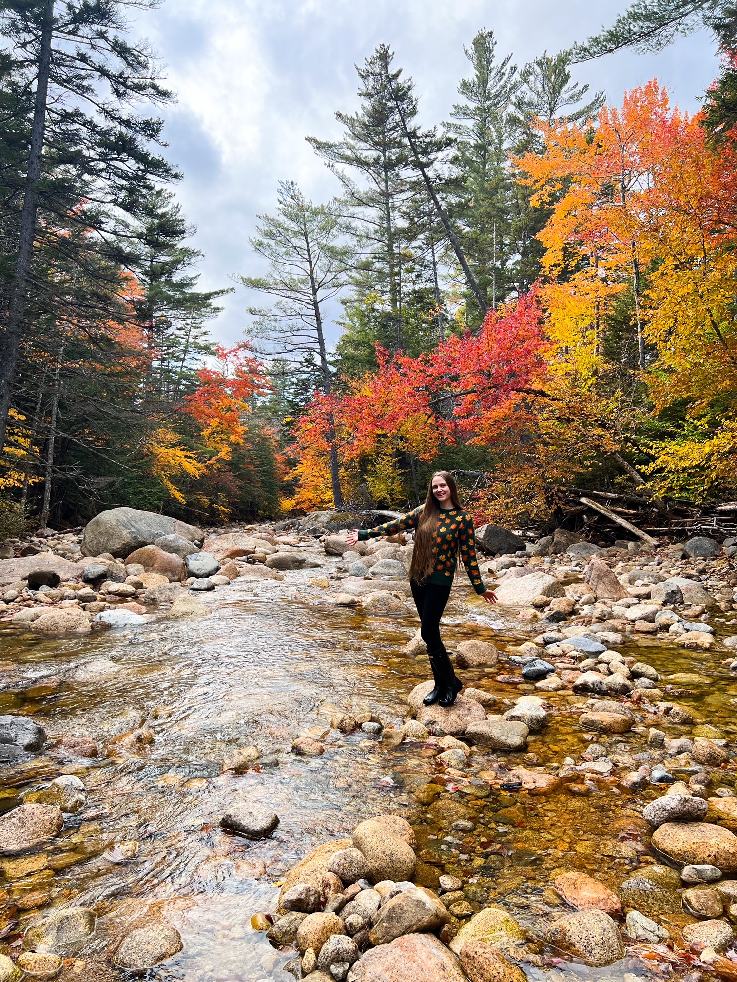 A woman in a fall-leaf sweater stands on a rock in the middle of a river, her hands spread out to show the largeness of the crossing and the color coordination her the trees and her sweater! 