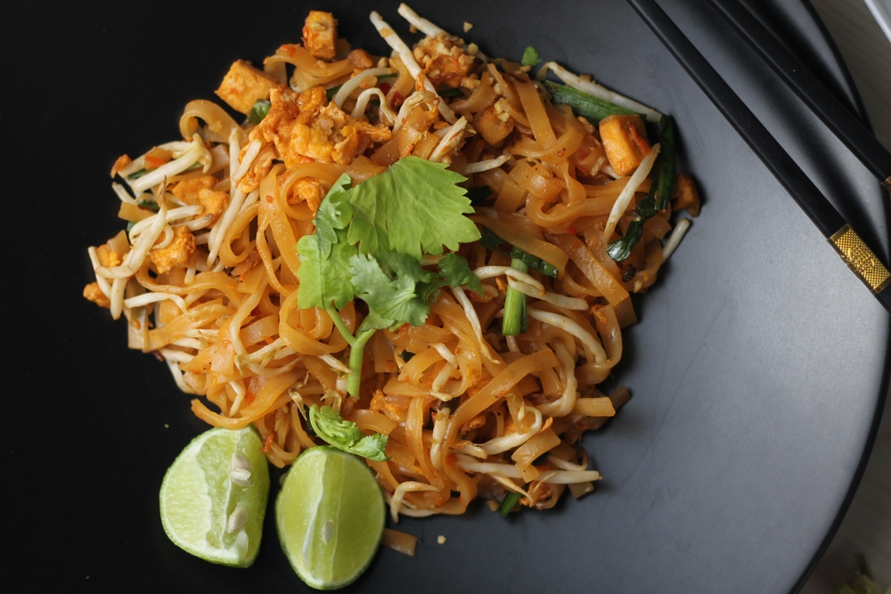 Pad Thai sits on a black plate with two limes, a bit of garnish and two chopsticks.