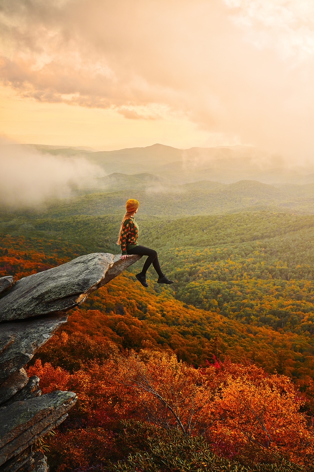 North Carolina is one of the prettiest states in the USA, especially in the fall, as seen by this photo that features a women in a yellow beanie sitting on the edge of a cliff and overlooking the endless trees and mountains in the distance that are bright will fall foliage. 